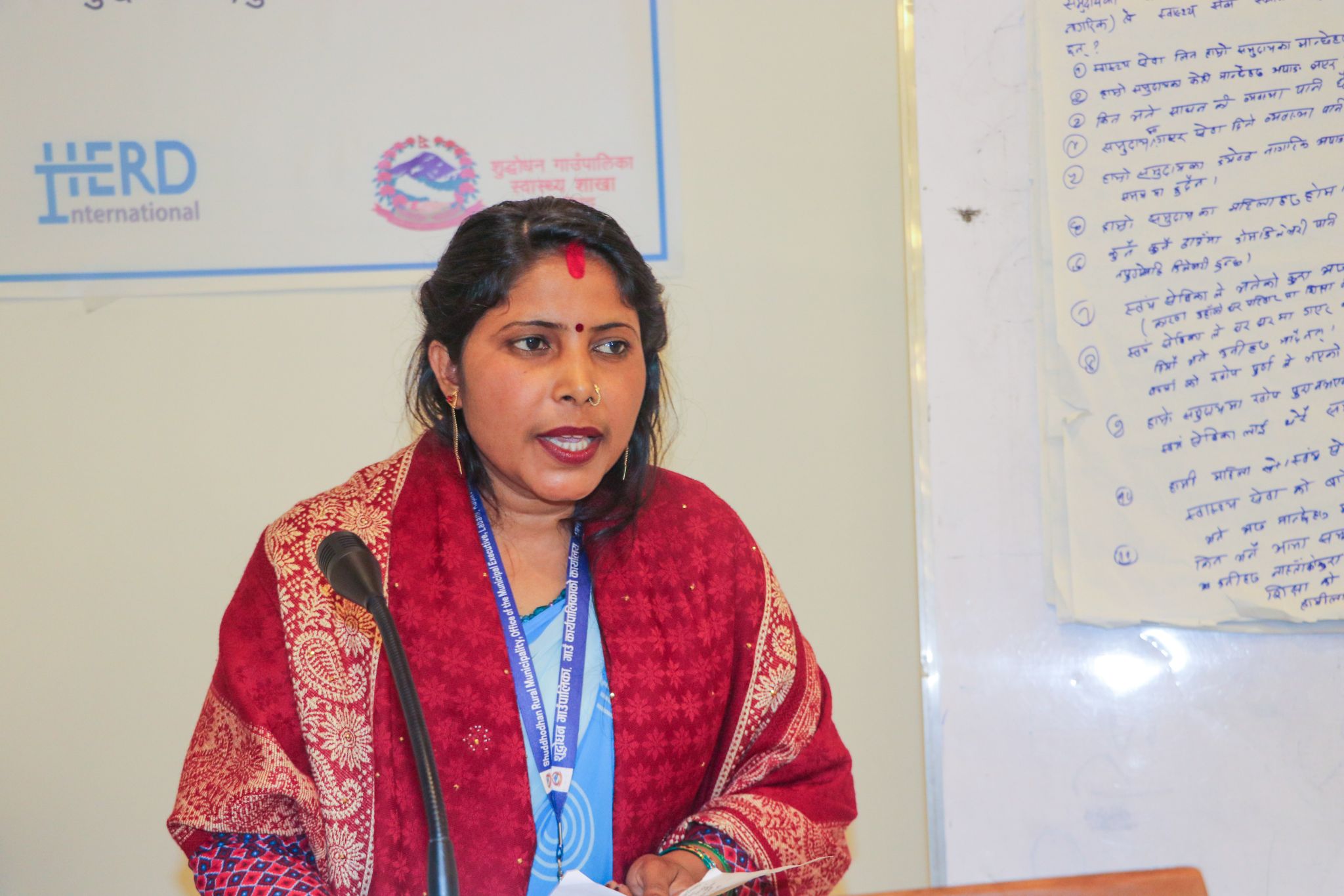 A Nepali woman in red and blue stands speaking to a room
