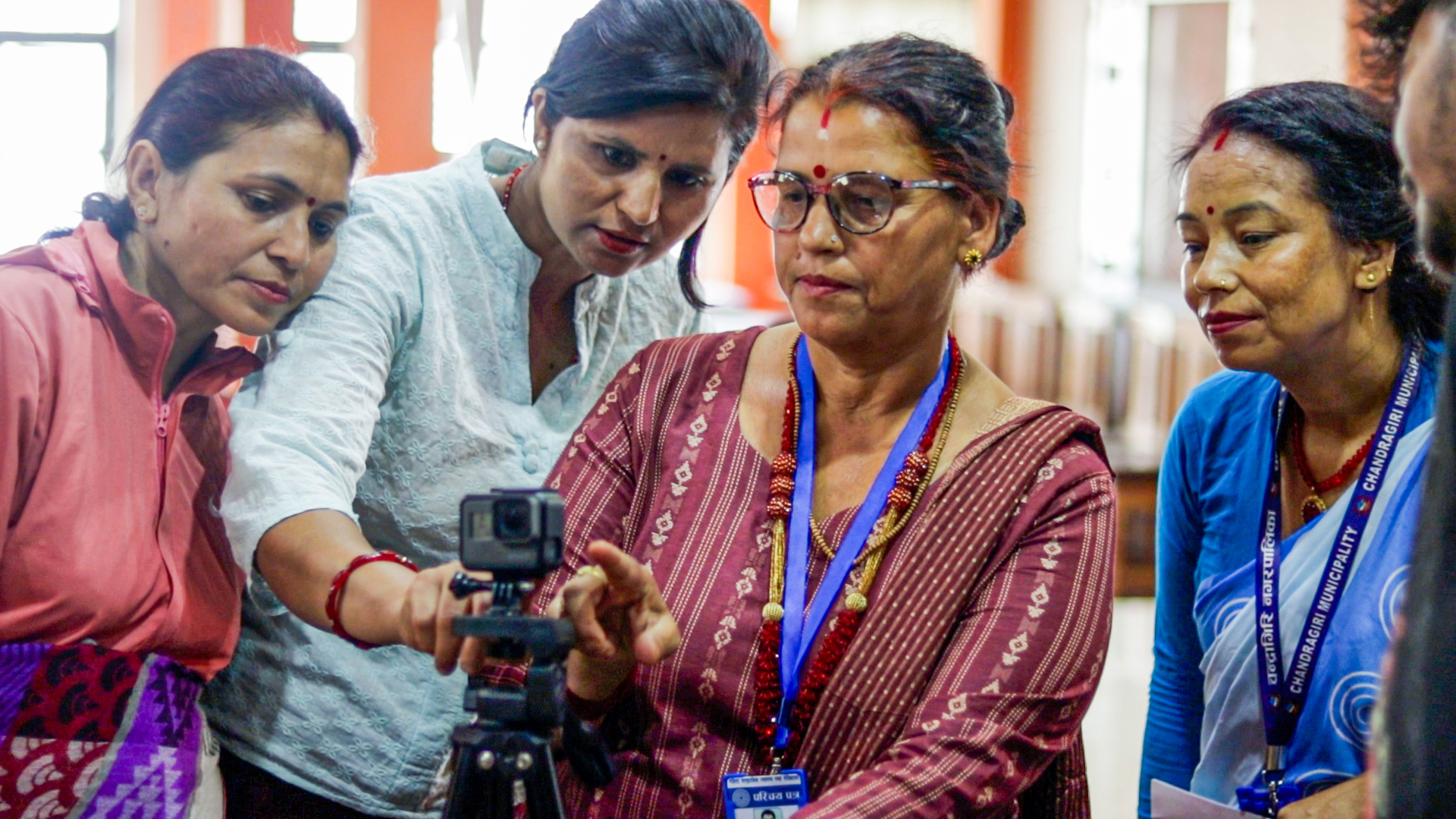 Four Nepali women gather around a camera on a tripod, looking at it intently