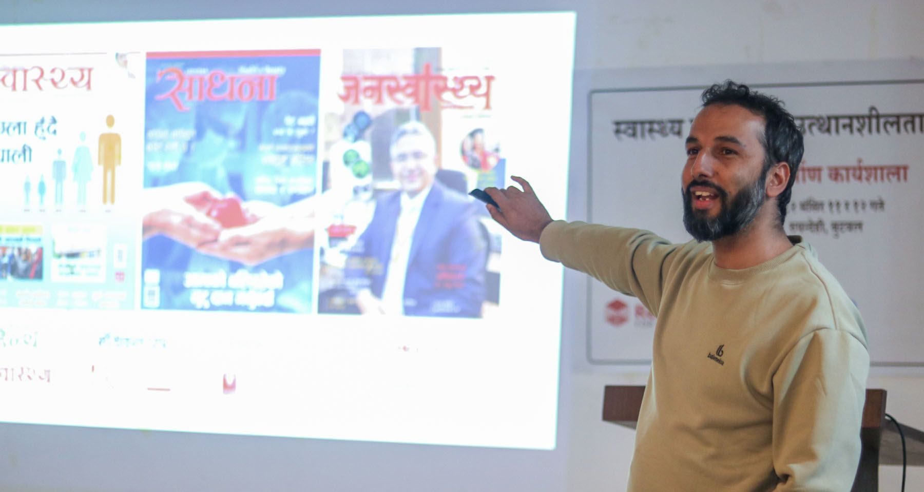A Nepali man stands speaking at the front of a room, pointing to a projection screen featuring the covers of publications