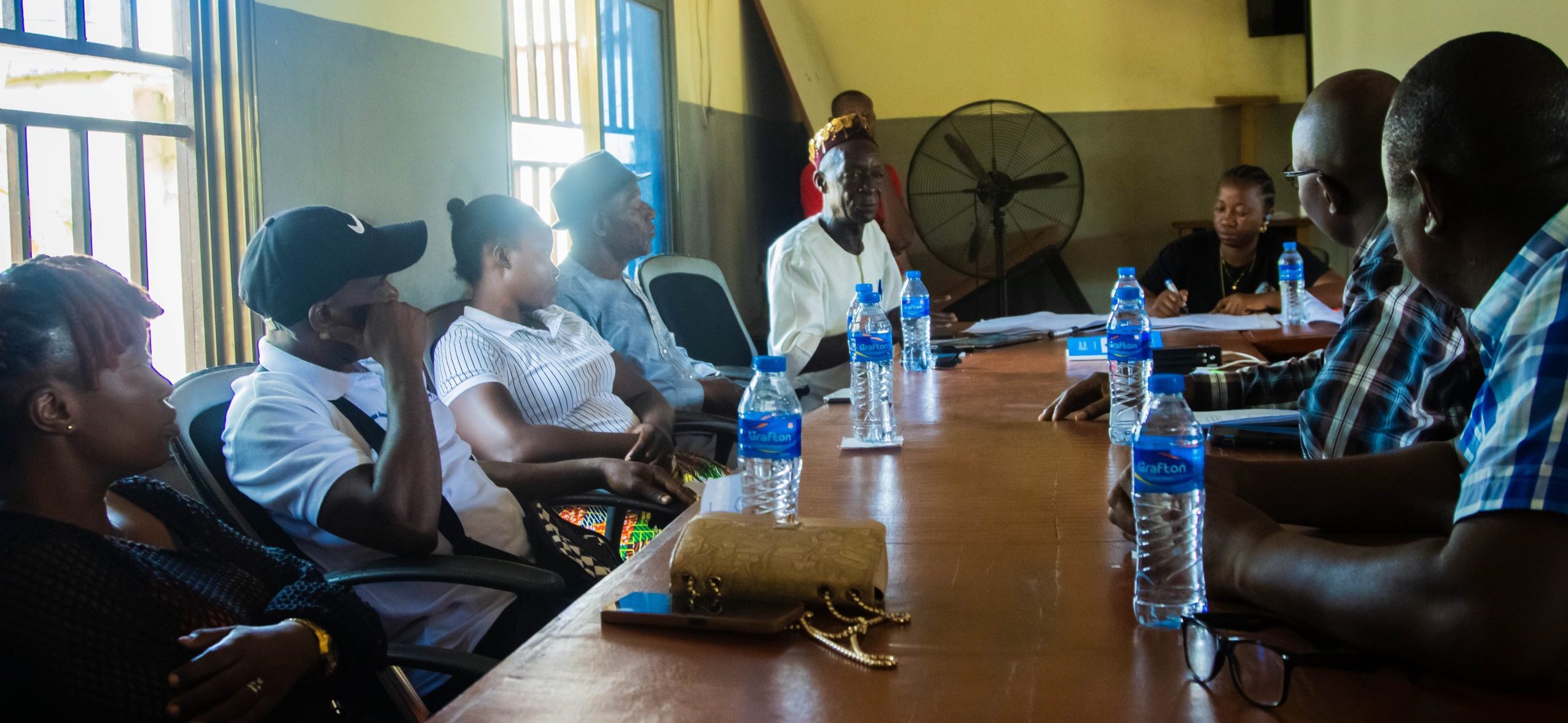 A group of Sierra leonian men and women sit around a table. A man with a hat (the chief) sits at the head of the table