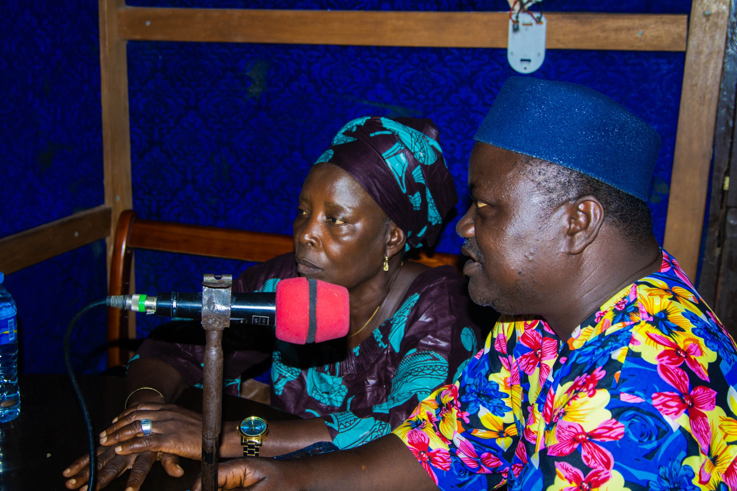A Sierra Leonian woman and man, both in brightly coloured clothes, sit speaking into a migraphone in a radio studio