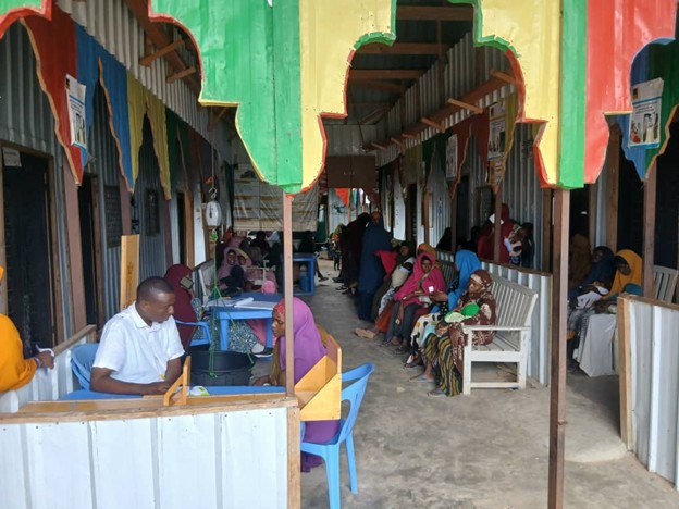 A long dark room with colourful awning and benches. Somalian people are sitting on benches waiting to be seen by a doctor