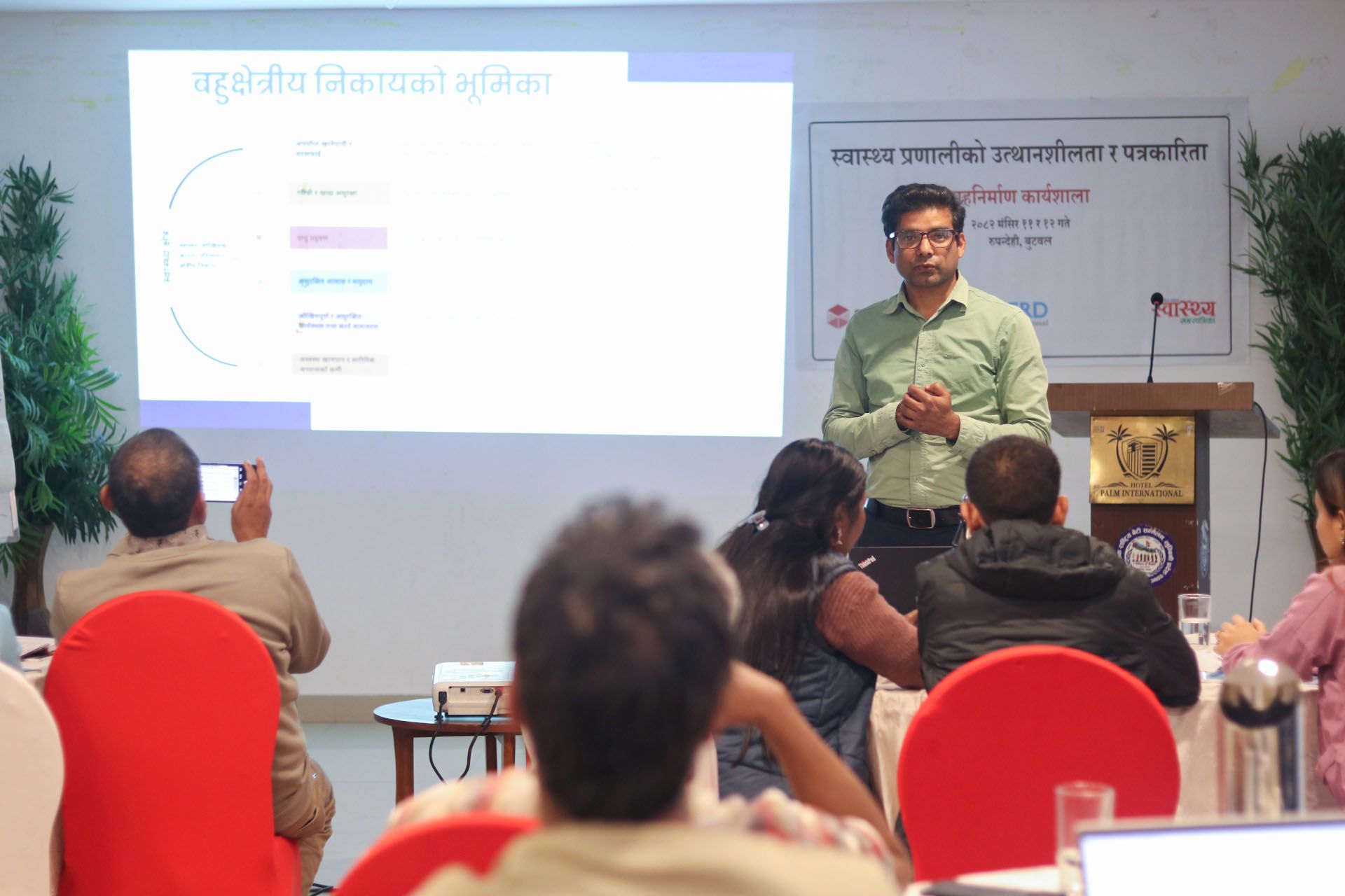 A Nepali man stands at the front of a room, speaking to those seated. Behind him is a projection screen