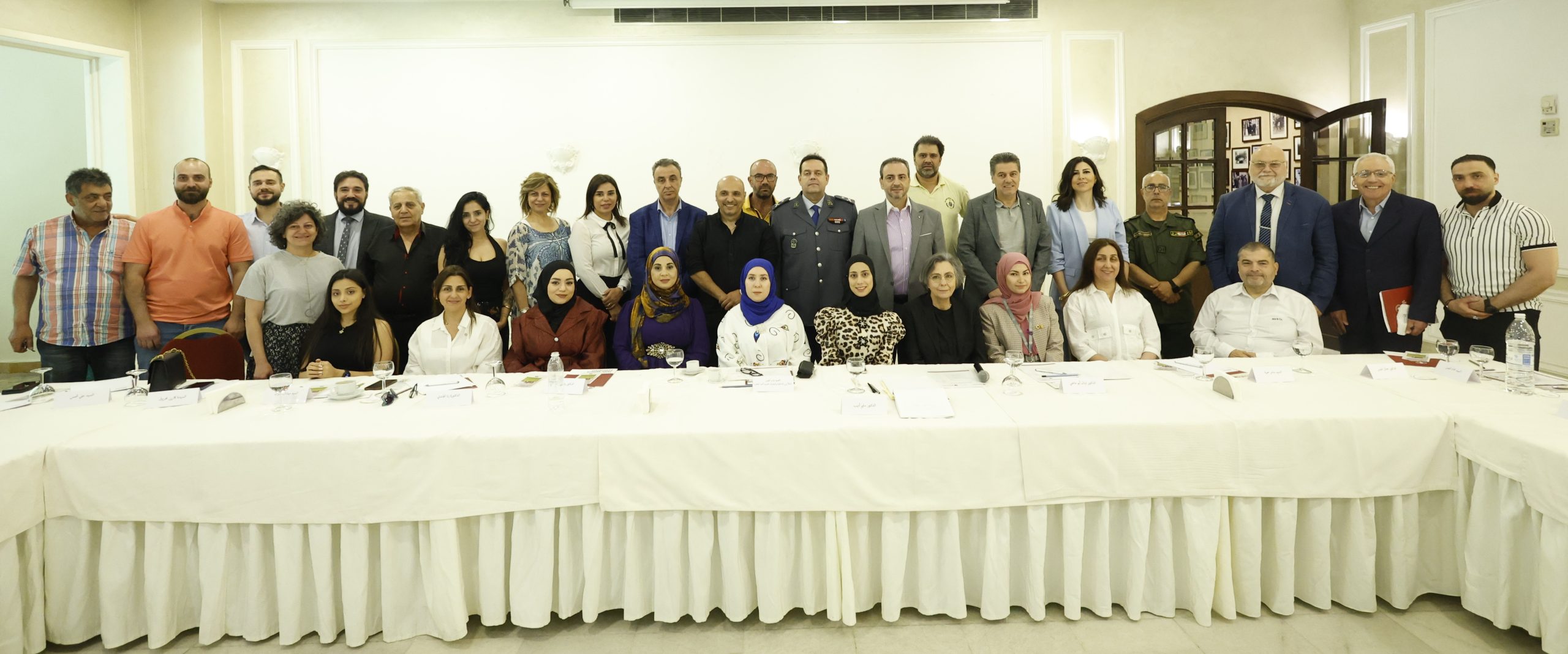 A long white table in a white room. Around the table stand and sit lots of Syrian and Lebanese people, all looking at the camera. Some are smiling,
