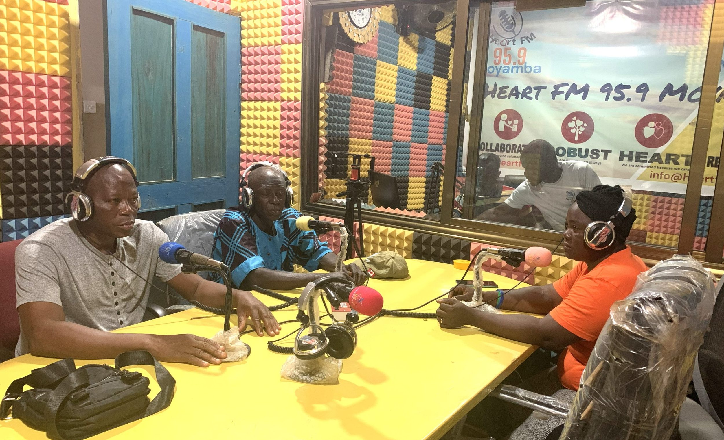 It's a radio studio in Sierra Leone. Three people sit around microphones talking, while in the booth a technician overseas the broadcast