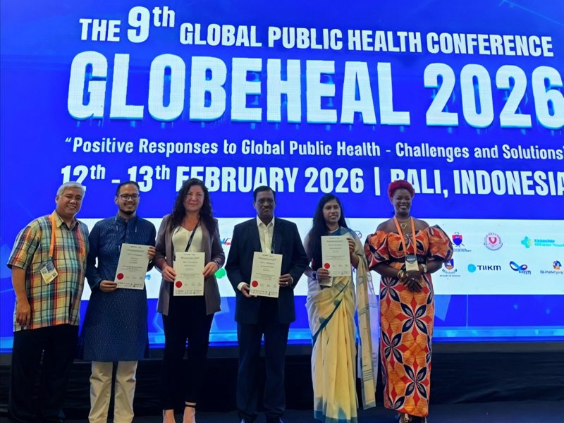 Six people standing, smiling on a stage in front of a large screen with 'Globeheal 2026' on it. Four of the people are holding certificates and smiling