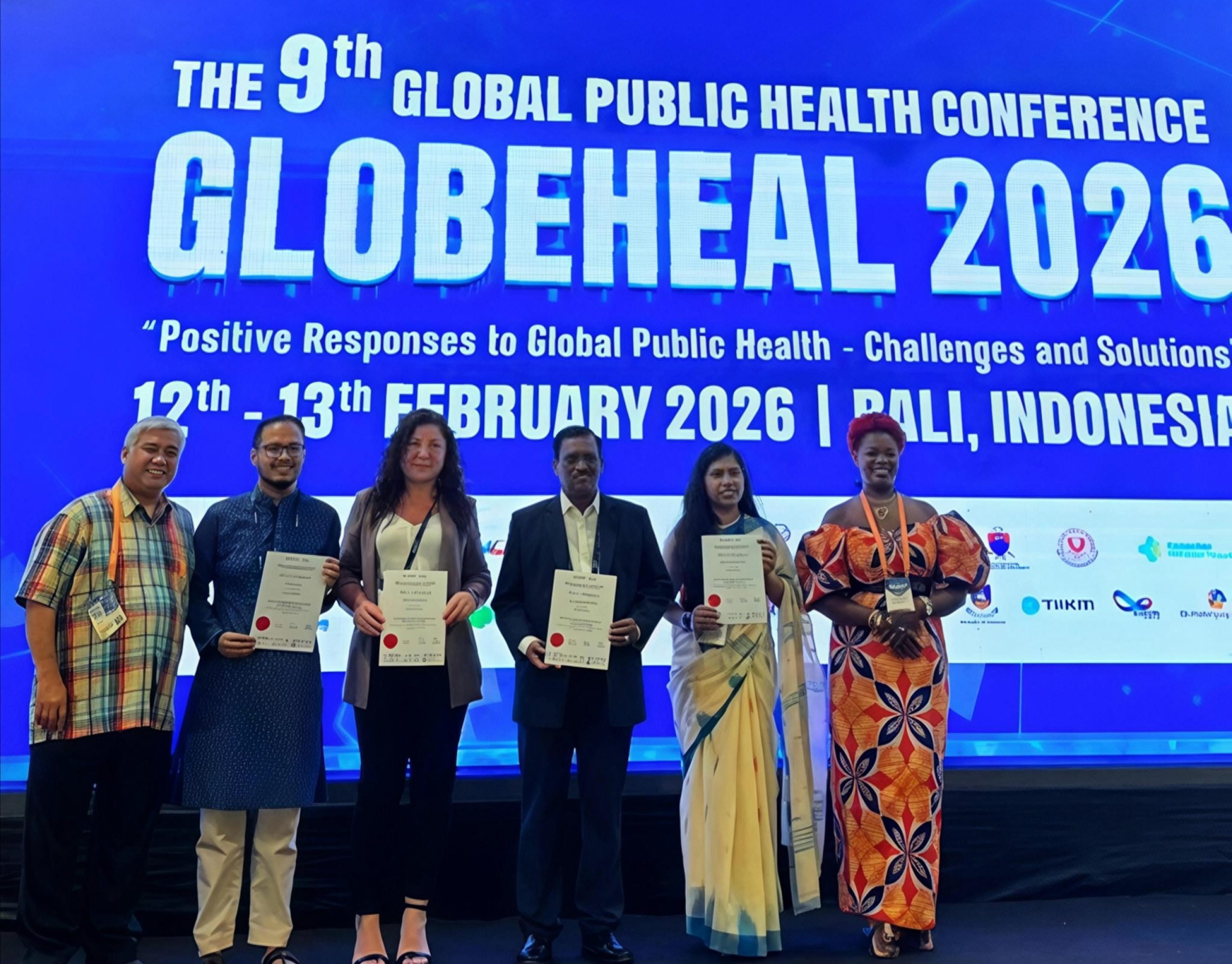 Six people standing, smiling on a stage in front of a large screen with 'Globeheal 2026' on it. Four of the people are holding certificates and smiling