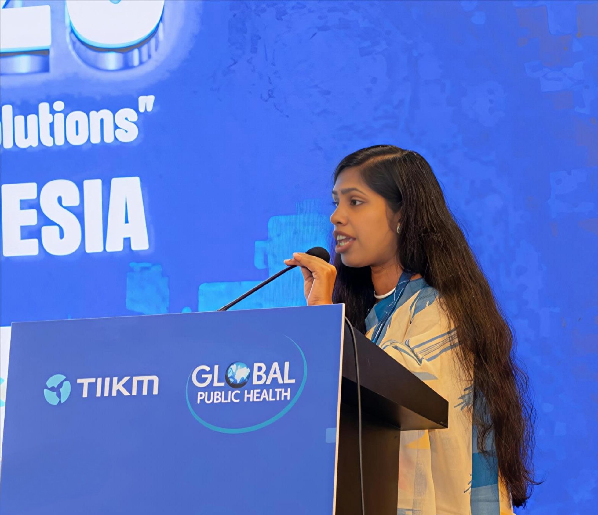 A Bangladeshi woman with long hair stands speaking into a microphone on a lectern on a stage. The lectern has 'Global Public Health' on it