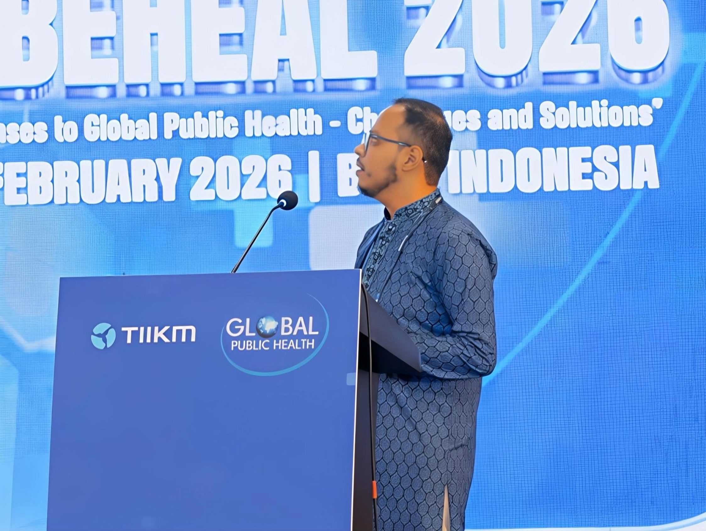 A Bangladeshi man wearing glasses speaks at a lectern on a stage