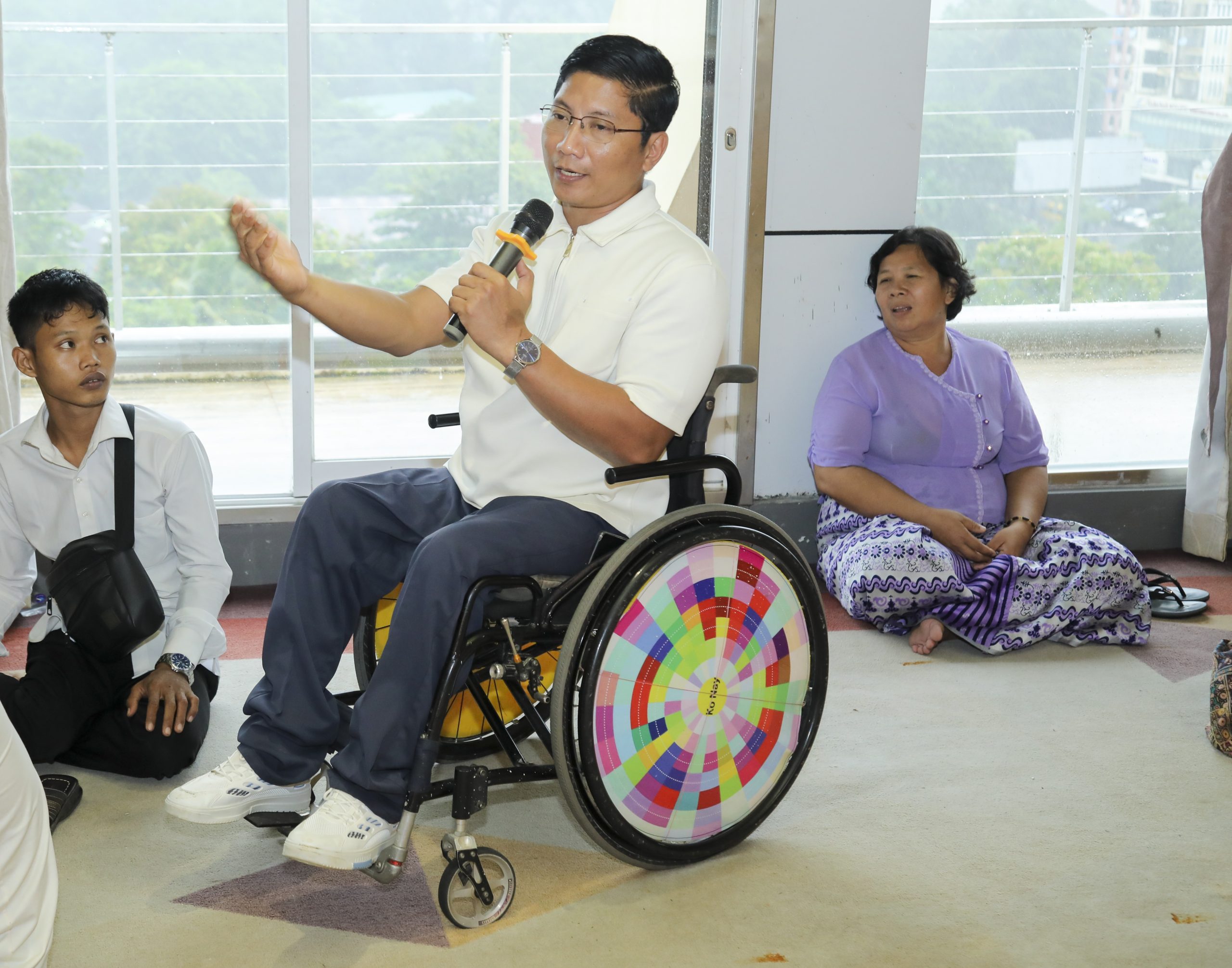 A Burmese man sitting in a wheelchair while speaking into a microphone. There are other people sitting on the floor around him listening