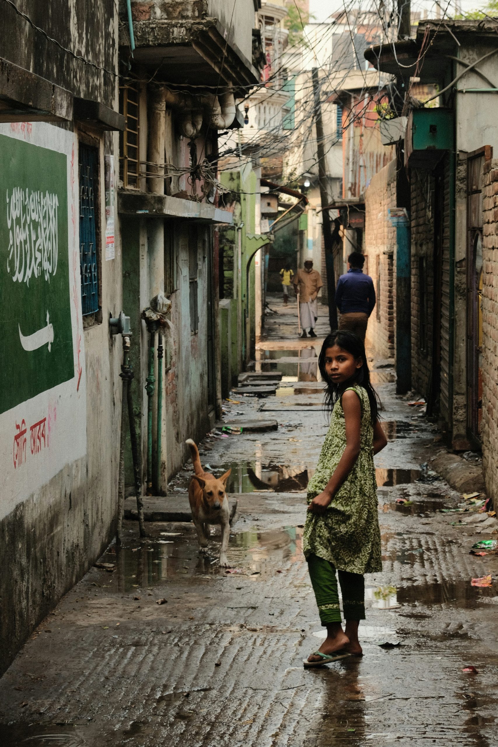 A girl is in a narrow alley way in Bangladesh, full of puddles. She looks back toward the camera