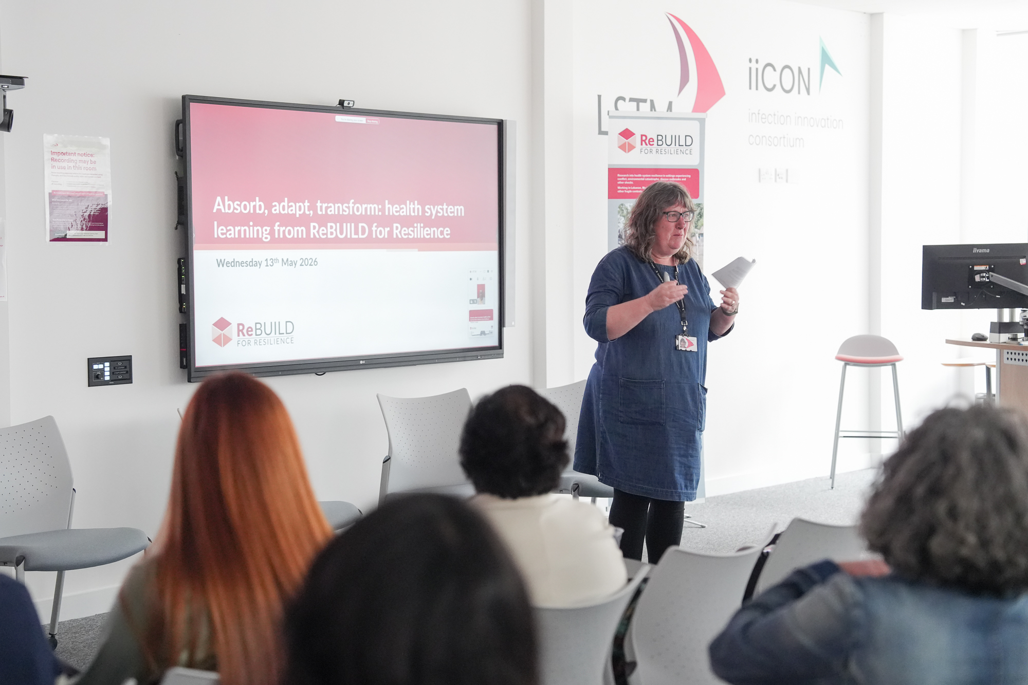 A woman stands speaking at the front of a room. There's a large screen behind her and we can see the backs of the audiences heads