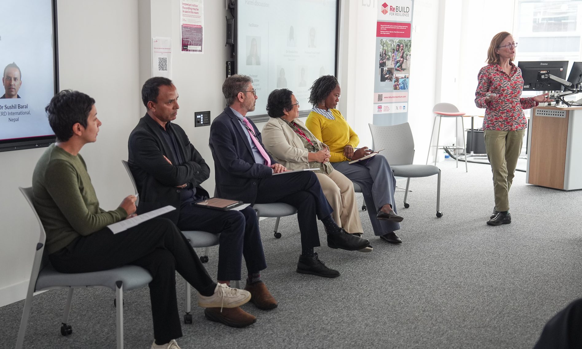 Five seated men and women at the front of a room. There's a projection screen behind them, a woman standing talking by a lectern next to them, and an audience just out of view