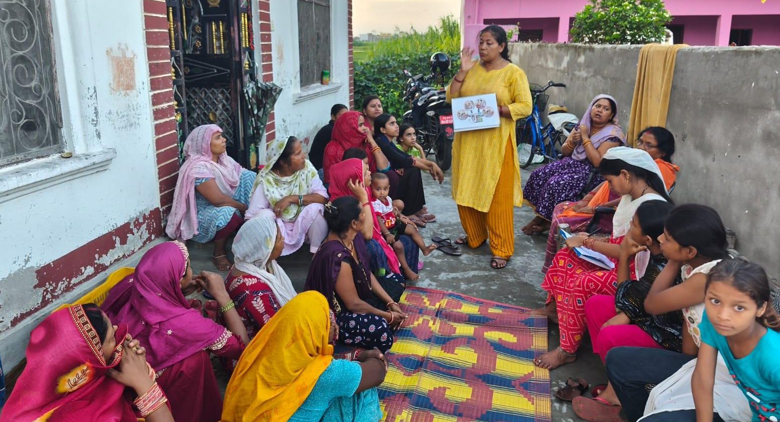 A female community health volunteer in Nepal stands speaking to a group of women and children sitting on the ground outside a building. They are all wearing brightly coloured clothes. She is holding a diagram with a health message
