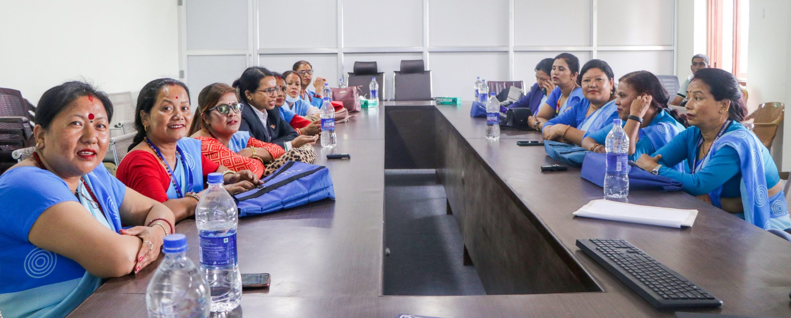 A long room with tables, at which are seated lots of Nepali ladies in blue saris. They are all looking at the camera